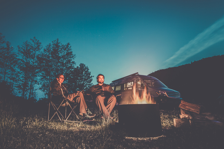 Couple Sitting Next to the Campfire with a Camper Van in the background. They are holding their hands.