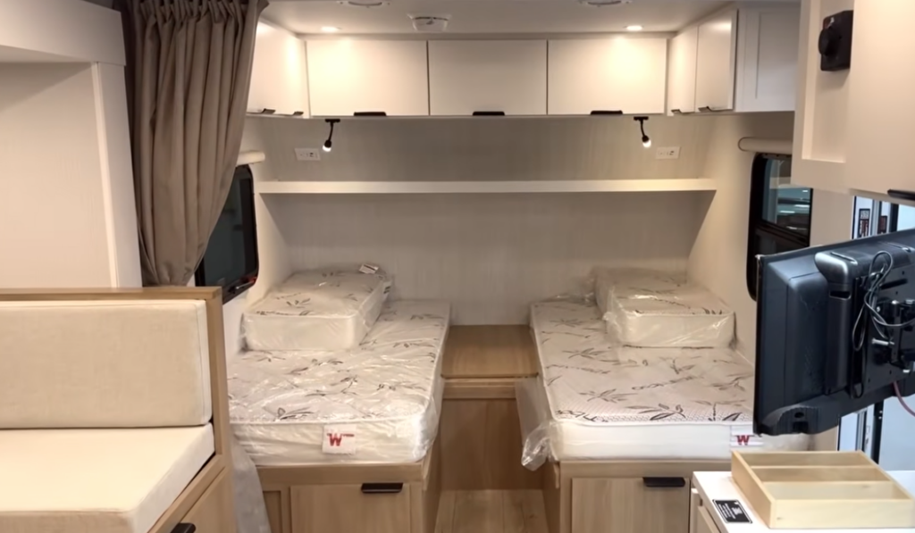 Modern RV rear bedroom featuring dual twin beds separated by a center nightstand, overhead storage cabinets, individual reading lights, and a bright residential-style interior designed for flexible sleeping arrangements and added comfort while traveling.