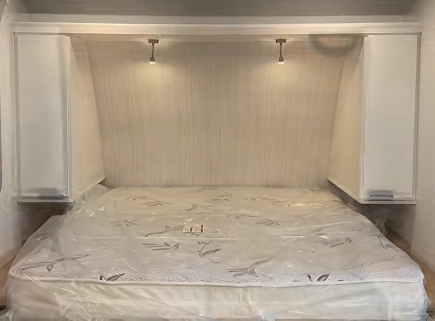 RV front bedroom featuring a residential-style queen bed, overhead reading lights, bedside wardrobes, and a clean modern design with bright white cabinetry and wood accents.