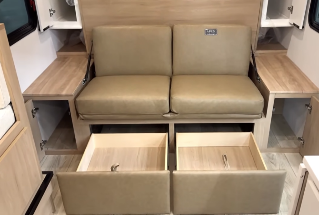 Compact RV sofa with dual storage drawers and side cabinets open, showcasing versatile seating, hidden storage solutions, and space-saving furniture design inside a lightweight travel trailer.