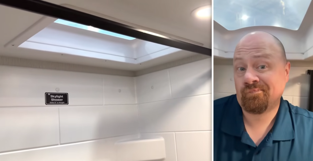 RV shower with a built-in skylight above the shower enclosure, providing additional headroom, natural light, and a more spacious feel for taller campers. A person stands beneath the skylight demonstrating the extra vertical clearance
