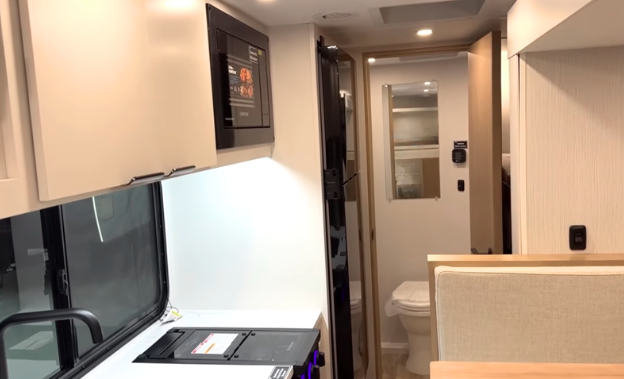 Modern travel trailer interior featuring a compact kitchen with solid-surface countertops, large refrigerator, microwave, and booth dinette. The view extends toward the rear bathroom, showcasing an efficient floor plan that maximizes living, cooking, and storage space in a lightweight RV.