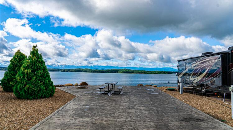 Premium waterfront RV campsite featuring a large concrete pad, picnic table, and luxury motorhome parked beside a scenic lake. The spacious site offers unobstructed water views, landscaped surroundings, and resort-style camping accommodations.