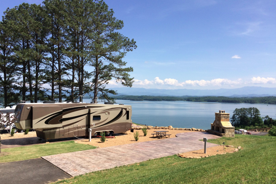 Luxury fifth wheel RV parked on a premium waterfront campsite overlooking a lake, featuring a spacious concrete pad, outdoor fireplace, picnic area, and panoramic water and mountain views at an upscale RV resort