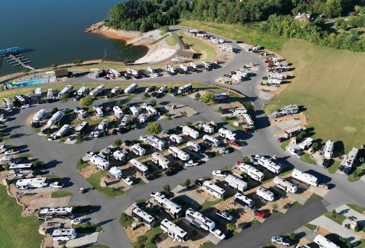 Aerial view of a large waterfront RV resort featuring dozens of RV campsites, paved roads, marina access, open green spaces, and direct access to a scenic lake. The resort offers a mix of seasonal and overnight RV sites with recreational amenities and beautiful water views
