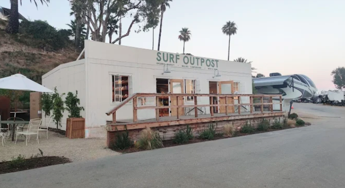 Entrance building at Surf Outpost RV Resort in Malibu, California, featuring a modern coastal design with outdoor seating, landscaped grounds, and RV sites nearby. The welcoming clubhouse serves as a central gathering space for guests enjoying a scenic beachfront camping experience