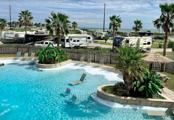 Resort-style RV campground pool featuring crystal-clear water, palm trees, in-water lounge chairs, and tropical landscaping. RV campsites are visible in the background, highlighting the blend of luxury resort amenities and coastal camping accommodations