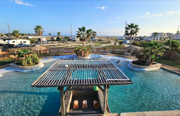 Resort-style swimming pool at a beachfront RV resort featuring a tropical island-inspired design, palm trees, shaded seating, and a central pavilion. The pool area offers guests a relaxing place to cool off while enjoying coastal views and upscale campground amenities