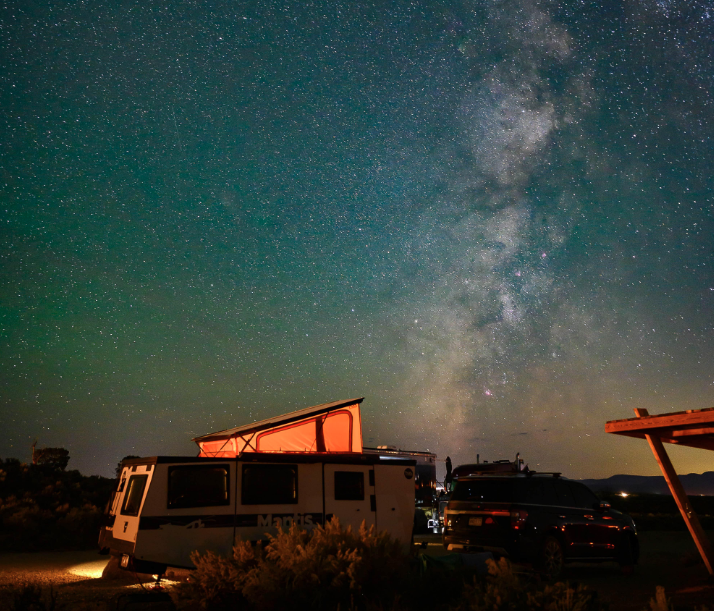 RV campsite under the Milky Way at a dark-sky campground, featuring an off-grid camper and tow vehicle illuminated by warm exterior lighting. Thousands of stars stretch across the night sky, creating a breathtaking stargazing experience for RV travelers and outdoor adventurers
