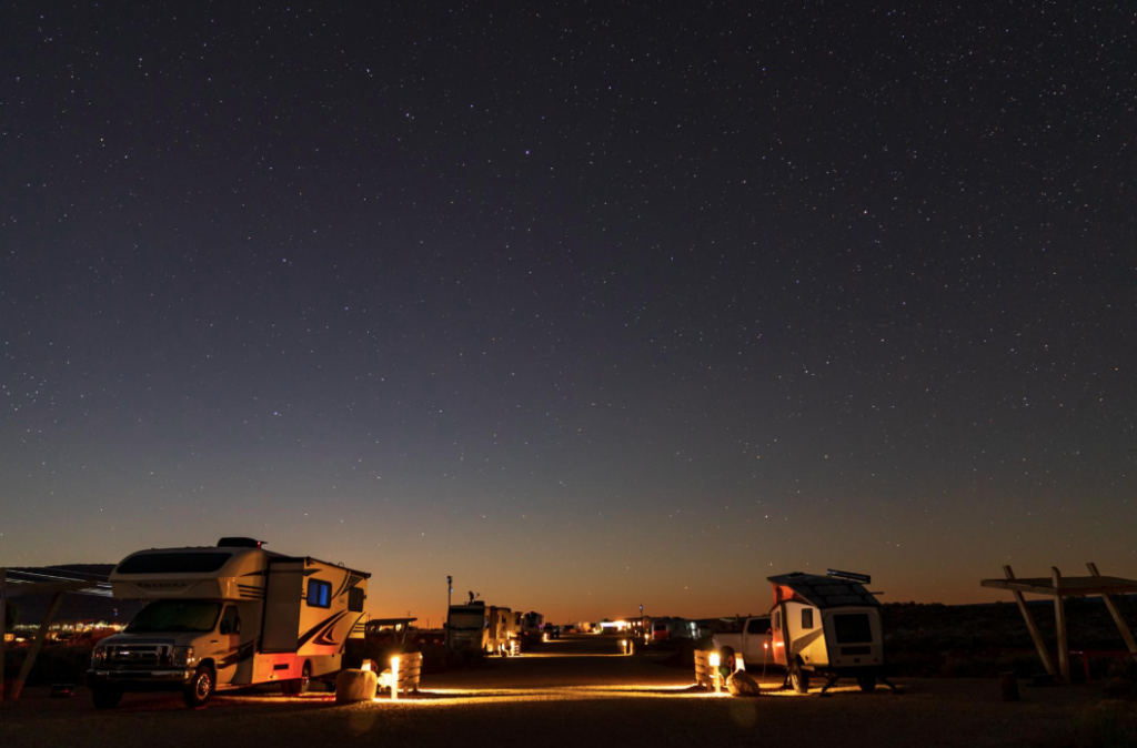 RVs and camper vans parked beneath a clear, star-filled night sky at a dark-sky RV campground. Soft campsite lighting illuminates the sites while the Milky Way and countless stars create an unforgettable stargazing experience for campers seeking peaceful nights and scenic desert views