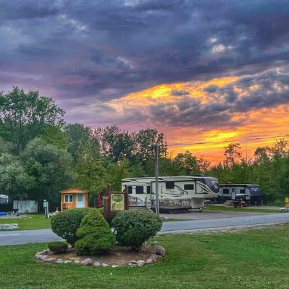 Scenic RV campground at sunset featuring luxury fifth wheel RVs, manicured landscaping, and spacious campsites surrounded by mature trees. Dramatic evening skies and colorful clouds create a peaceful atmosphere for campers enjoying a relaxing stay at a full-service RV resort