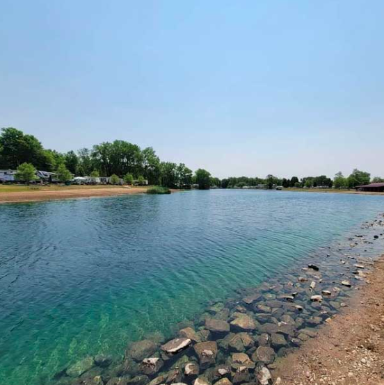 Crystal-clear lake at a waterfront RV resort featuring a rocky shoreline, sandy beach areas, and scenic campsites nestled among mature trees. The calm blue water provides opportunities for swimming, kayaking, fishing, and relaxing outdoor recreation during an RV getaway