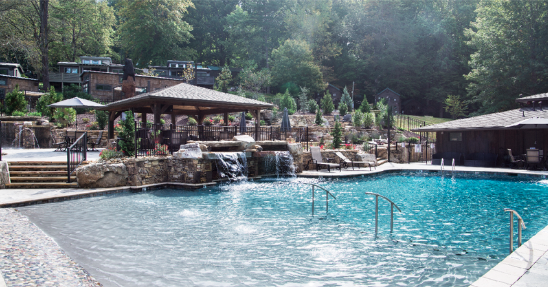 Resort-style swimming pool at Little Arrow Outdoor Resort in Townsend, Tennessee, featuring cascading waterfalls, stone landscaping, shaded gathering areas, and a peaceful mountain setting. Surrounded by wooded hillsides and luxury accommodations, the pool offers guests a relaxing retreat near Great Smoky Mountains National Park