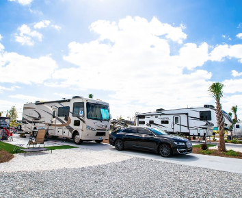 Luxury RV resort campsite featuring a Class A motorhome, fifth wheel RV, paved parking area, and tropical landscaping with palm trees. The spacious full-hookup site offers modern amenities, resort-style accommodations, and a welcoming atmosphere for extended RV stays and vacation getaways