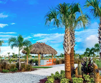 Tropical-style amenities area at a luxury RV resort featuring palm trees, lush landscaping, a thatched-roof pavilion, and colorful outdoor seating. The inviting outdoor space creates a relaxing coastal atmosphere where guests can gather, unwind, and enjoy resort-style camping accommodations