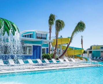 Resort-style swimming pool complex featuring a large waterslide, fountains, palm trees, and rows of lounge chairs at a luxury RV resort. The family-friendly aquatic area offers guests a fun and relaxing vacation experience with upscale amenities and a tropical atmosphere