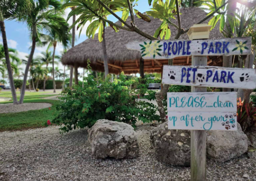 Pet-friendly RV resort featuring a designated dog park area surrounded by tropical landscaping, palm trees, and resort amenities. A rustic sign directs guests to the pet park, highlighting the campground’s welcoming atmosphere for RV travelers camping with their dogs