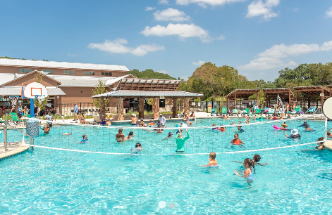 Family-friendly resort swimming pool filled with guests enjoying water activities, swimming, and recreation. The spacious aquatic center features resort-style amenities, shaded gathering areas, and plenty of room for families to relax and have fun during their RV camping vacation
