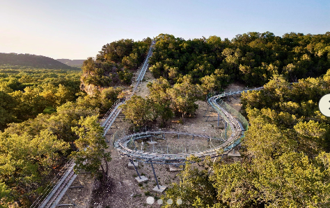 Mountain coaster attraction winding through a forested hillside at an outdoor adventure resort. The elevated track curves through scenic woodland terrain, offering guests a thrilling ride and unique recreational experience surrounded by natural beauty and panoramic views