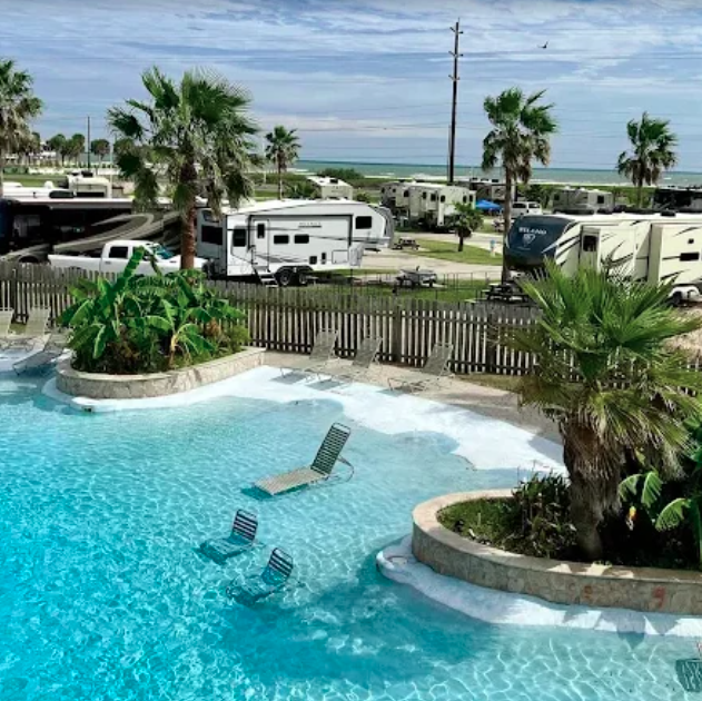 Resort-style swimming pool with in-water lounge chairs, tropical landscaping, and palm trees overlooking a coastal RV resort. Luxury RVs and full-hookup campsites surround the pool area, creating a relaxing vacation atmosphere that combines beachfront camping with upscale resort amenities