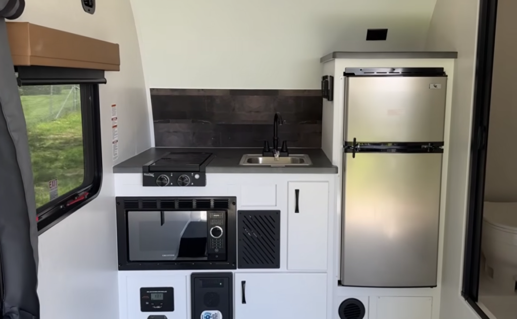 Compact RV kitchen inside the Modern Buggy Hopper travel trailer featuring a refrigerator, microwave, sink, induction cooktop, and efficient storage cabinets. The thoughtfully designed galley maximizes functionality in a lightweight, adventure-ready camper built for comfortable travel and easy towing