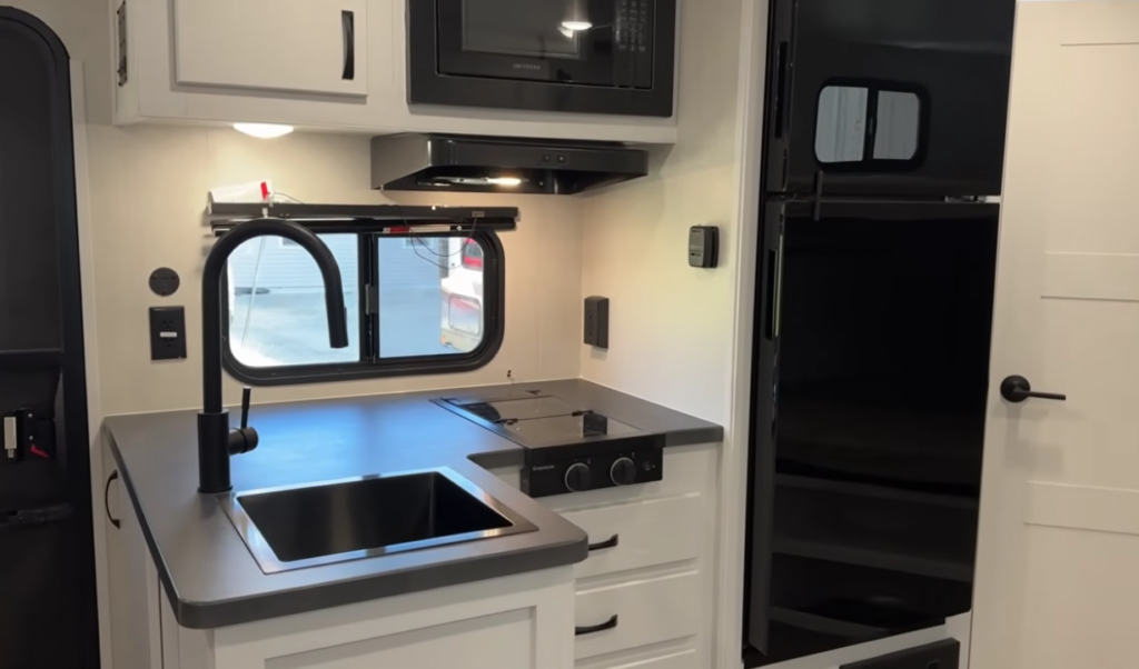 Modern kitchen inside the Modern Buggy Hopper travel trailer featuring a deep stainless steel sink, high-rise faucet, two-burner cooktop, microwave, residential-style refrigerator, and ample countertop workspace. The contemporary galley design maximizes storage, functionality, and convenience, providing everything needed for comfortable camping in a lightweight adventure-ready RV