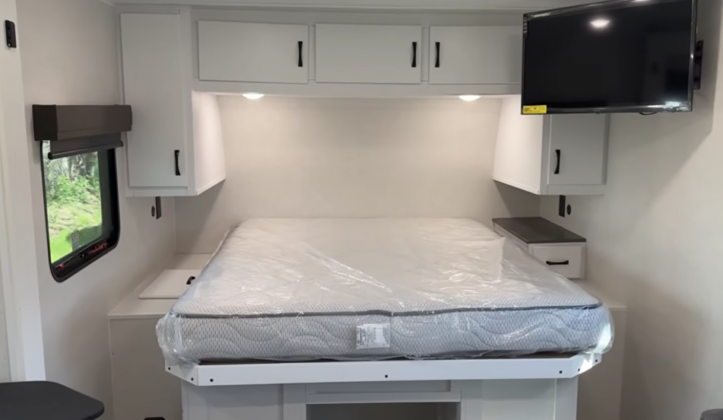 Private front bedroom inside the Modern Buggy Hopper travel trailer featuring a residential queen-size bed, overhead storage cabinets, bedside wardrobes, built-in nightstands, and a mounted TV for entertainment. The bright, modern design maximizes comfort and storage while providing a cozy retreat after a day of outdoor adventure
