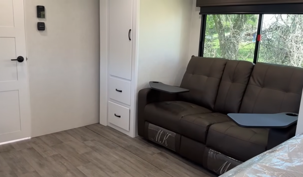 Comfortable living area inside the Modern Buggy Hopper travel trailer featuring a plush tri-fold sofa with integrated cupholder armrests, large panoramic window, built-in storage drawers, and an open floorplan. The residential-style seating provides a relaxing space to unwind, entertain, or convert for additional sleeping accommodations in this lightweight, adventure-ready RV
