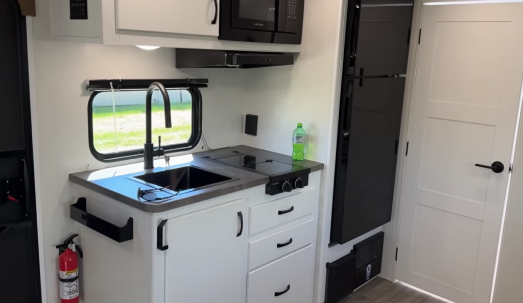 Modern RV kitchen featuring a deep farmhouse-style sink, microwave, two-burner cooktop, residential refrigerator, and ample storage cabinetry. The bright, contemporary design maximizes functionality and comfort, making meal preparation easy in this compact travel trailer interior.