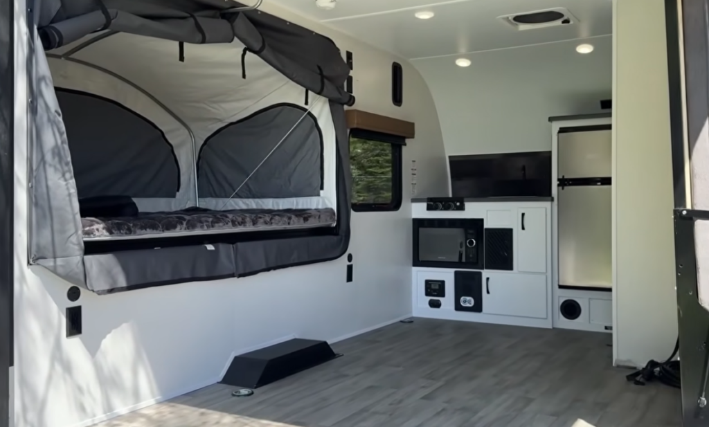 Open interior of the Modern Buggy Hopper travel trailer featuring a fold-out canvas sleeping pod, spacious multi-purpose living area, microwave, refrigerator, and compact kitchen storage. The innovative design combines the comfort of a hard-sided travel trailer with the open-air camping experience of a tent camper, creating a lightweight and versatile RV for outdoor adventures