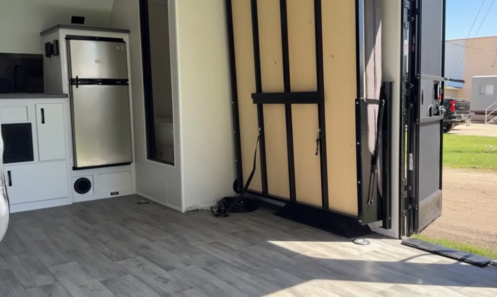 Open-concept interior of the Modern Buggy Hopper travel trailer showcasing its versatile cargo and living space. The rear wall-mounted fold-away bed creates a spacious multi-purpose area for hauling gear, relaxing, or customizing the layout, while the compact kitchen, refrigerator, and private bathroom remain fully accessible. This innovative design blends toy-hauler flexibility with the comfort of a lightweight travel trailer built for adventure