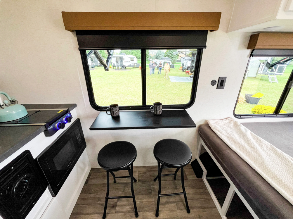 Compact dining and living area inside the Modern Buggy Hopper travel trailer featuring a window-side bar table with seating for two, large panoramic windows, a kitchenette with microwave and cooktop, and a convertible sleeping/lounge space. The efficient layout maximizes comfort, functionality, and campsite views in a lightweight adventure-ready RV