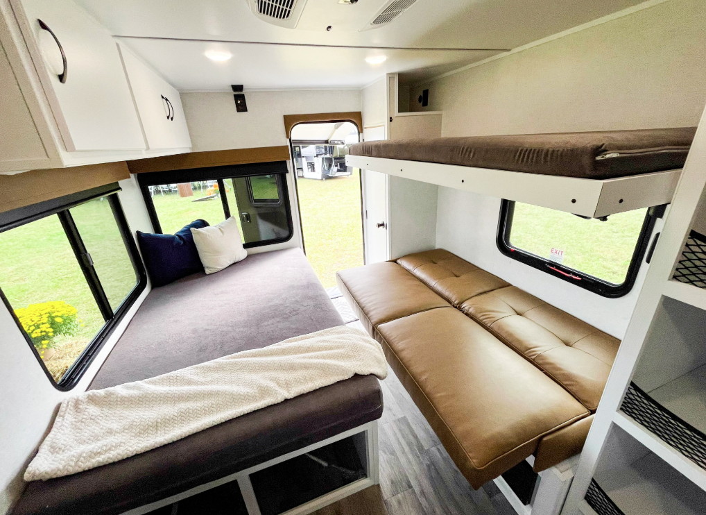 Multi-purpose living and sleeping area inside the Modern Buggy Hopper travel trailer featuring a large convertible bed, jackknife sofa, overhead bunk, panoramic windows, and rear entry door access. The flexible interior layout maximizes sleeping capacity, storage, and comfort while maintaining the compact footprint of this lightweight adventure-focused RV
