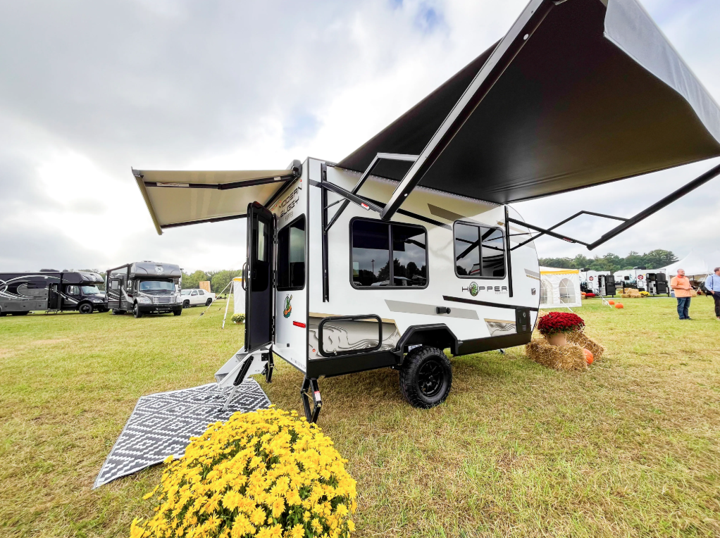 odern Buggy Hopper travel trailer displayed at an RV show featuring dual oversized power awnings, rugged off-road tires, a compact lightweight design, and large tinted windows.