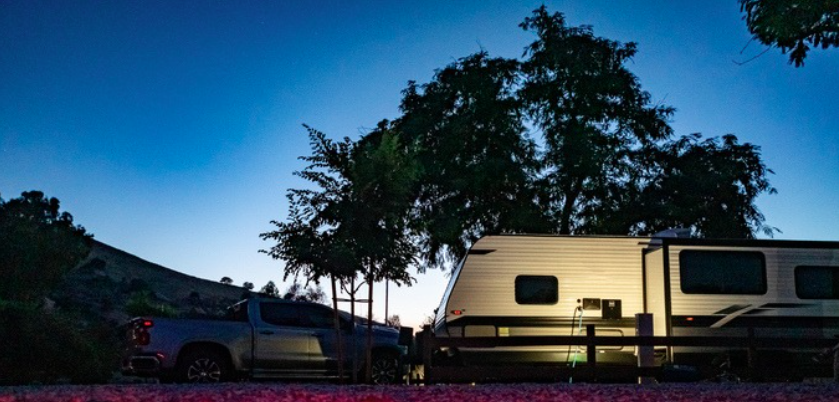 Travel trailer parked at a campsite during dusk with soft exterior lighting illuminating the RV and tow vehicle beneath a clear evening sky. The peaceful campground setting highlights the comfort, convenience, and freedom of RV travel, showcasing a relaxing overnight camping experience surrounded by nature.