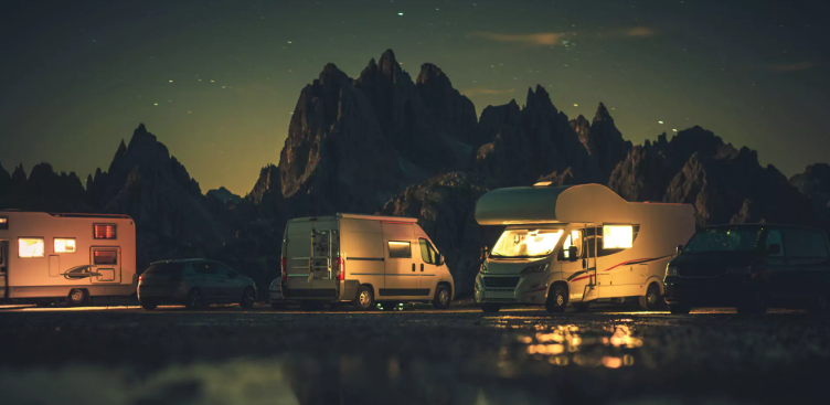 Motorhomes and camper vans parked beneath a breathtaking star-filled night sky with dramatic mountain peaks in the background. Warm interior lights glow from the RVs, creating a peaceful campground atmosphere that captures the beauty of overnight RV travel, stargazing, and unforgettable outdoor adventures in scenic destinations.