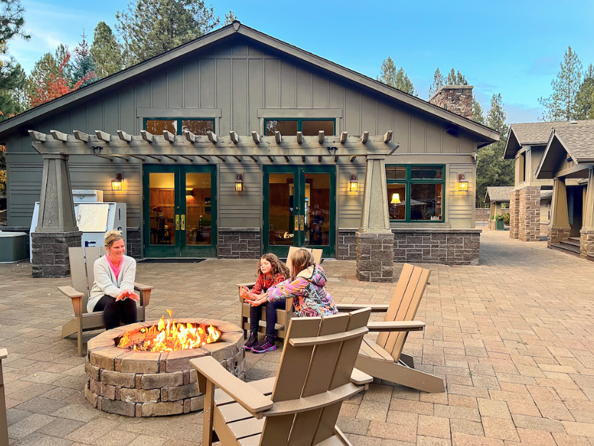 Campground clubhouse with a large outdoor fire pit surrounded by Adirondack chairs where a family relaxes together. The welcoming community gathering space features a lodge-style building, spacious patio, and cozy fire pit, creating the perfect place for RV campers to unwind, connect with friends and family, and enjoy evenings at a full-service RV resort.