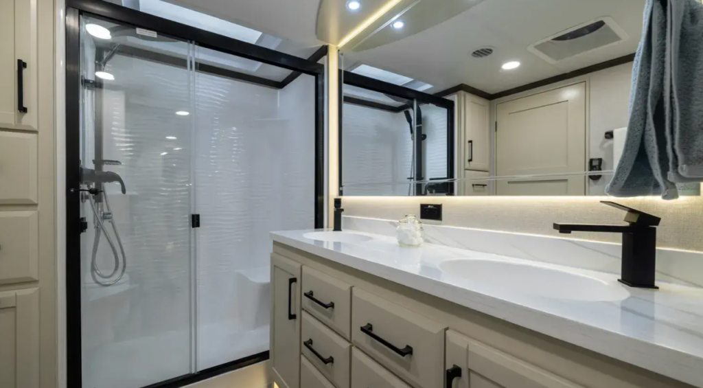 Luxury fifth wheel master bathroom featuring a dual-sink vanity with solid-surface countertops, illuminated residential-style mirror, abundant drawer storage, and a spacious glass-enclosed shower with built-in seating. Designed for full-time RV living, the premium bathroom delivers residential comfort, modern finishes, and ample space for extended camping and luxury travel.