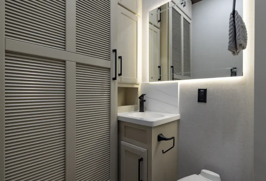 Luxury fifth wheel bathroom featuring a modern vanity with an illuminated mirror, solid-surface sink, residential-style cabinetry, and a spacious linen closet with louvered doors. Designed for full-time RV living, the upscale bathroom offers generous storage, contemporary finishes, and residential comfort for extended camping and travel.