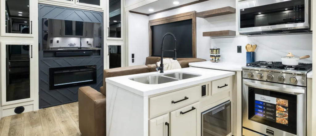 Luxury fifth wheel kitchen featuring a large residential island with stainless steel sink, solid-surface countertops, premium residential gas range, microwave, and modern cabinetry. The open-concept living space includes high-end finishes, an electric fireplace, comfortable theater seating, and abundant storage, showcasing the upscale residential comfort and craftsmanship found in a luxury RV designed for full-time living and extended travel.