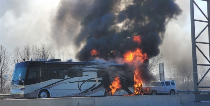 Motorhome engulfed in flames on the side of a highway with thick black smoke billowing into the air.