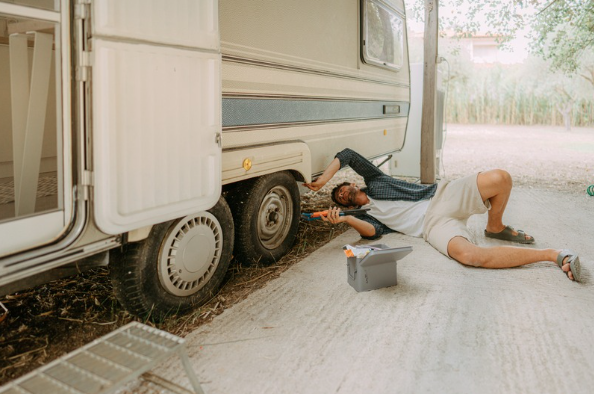 RVer lying beneath a travel trailer performing routine maintenance or inspecting the suspension and running gear with a toolbox nearby.