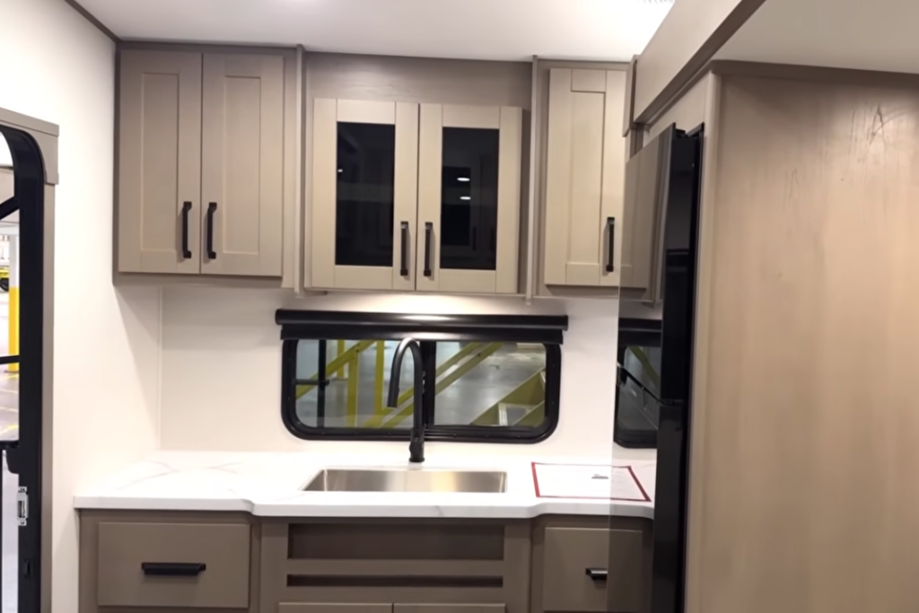 Residential-style RV kitchen featuring a large stainless-steel sink, solid-surface countertops, abundant overhead cabinetry, pantry storage, and a large window that brings in natural light.