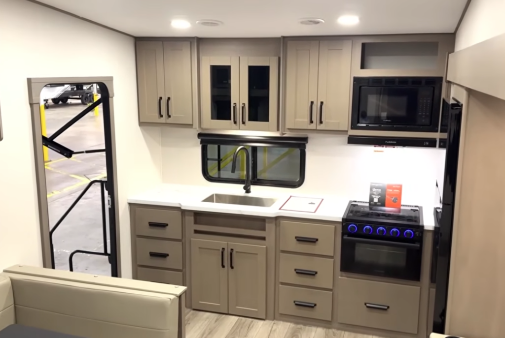 Modern RV kitchen featuring residential-style cabinetry, solid-surface countertops, a large stainless-steel sink, three-burner gas range with oven, microwave, generous drawer storage, and a spacious refrigerator.