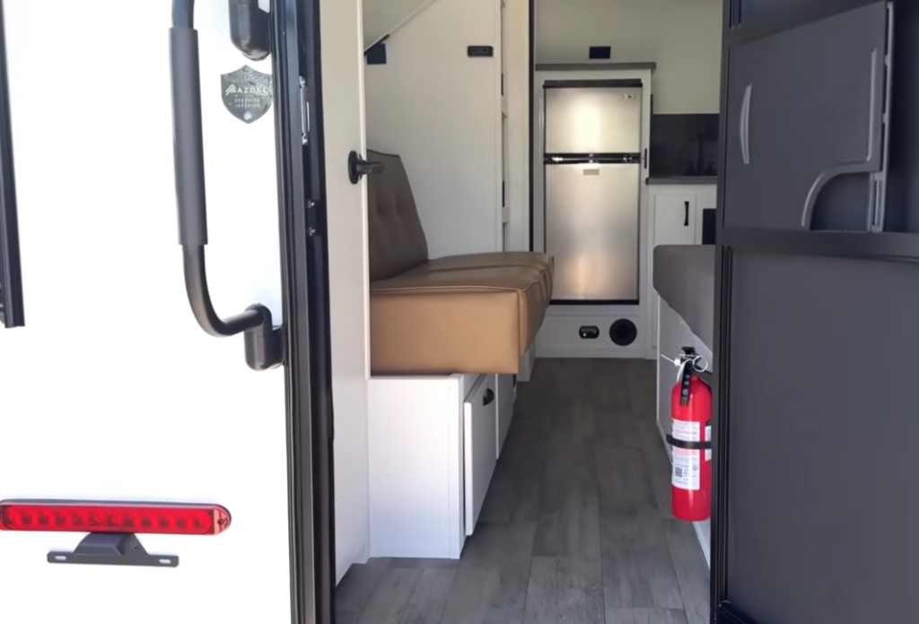 View through the rear entry door of the Modern Buggy Hopper travel trailer, showcasing its bright, open-concept interior with a convertible sofa, spacious floor space, residential refrigerator, compact kitchen, and convenient fire extinguisher near the entrance.