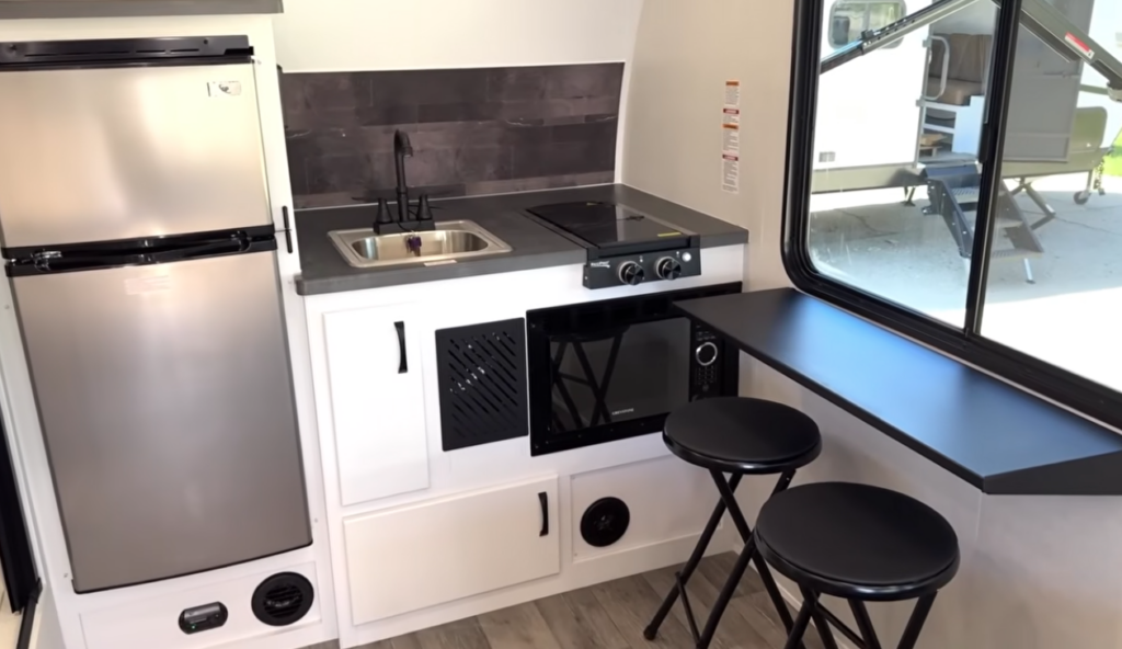 Compact kitchen inside the Modern Buggy Hopper travel trailer featuring a two-burner cooktop, stainless-steel sink, microwave, residential-style refrigerator, and modern cabinetry.