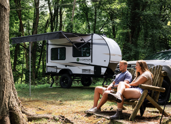 Couple relaxing at a wooded campsite beside the Modern Buggy Hopper travel trailer with the power awning extended.