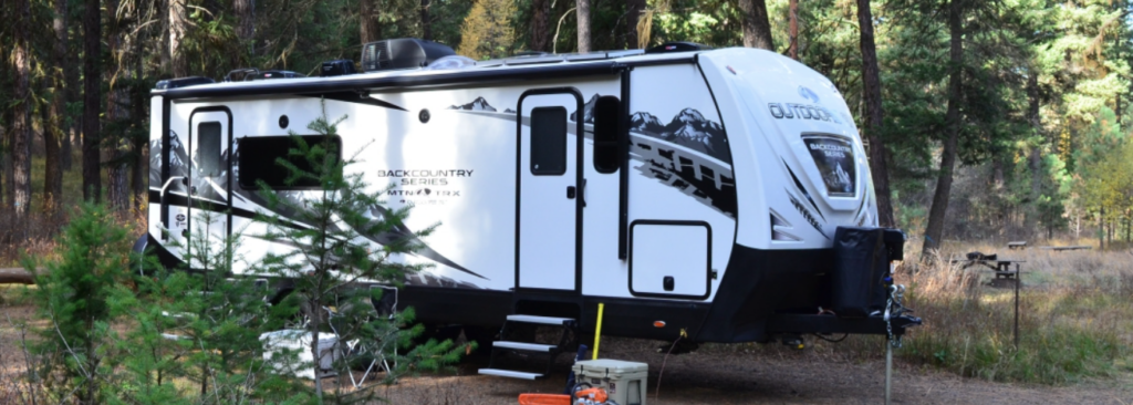 Outdoors RV Back Country Series travel trailer set up at a forest campground surrounded by tall pine trees, showcasing its rugged, off-grid-ready design.