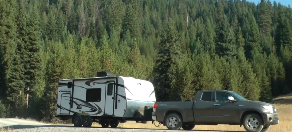 Outdoors RV travel trailer being towed by a full-size pickup truck through a scenic mountain landscape surrounded by evergreen forests.