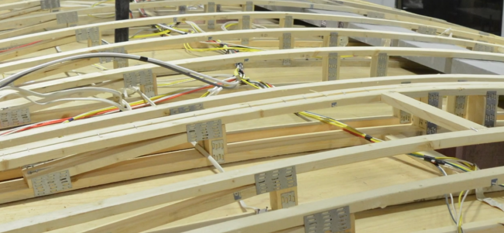 Exposed roof framing of an Outdoors RV travel trailer during the manufacturing process, showcasing the arched wood roof trusses, integrated wiring, and heavy-duty construction beneath the finished roof.